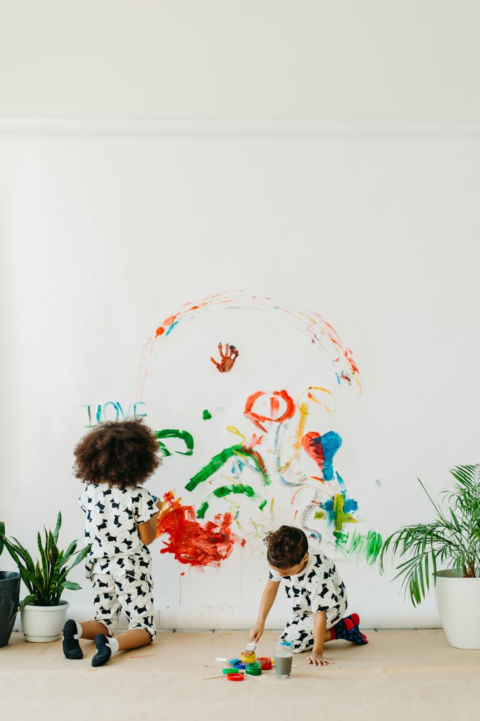 Two children create vibrant wall art with paints indoors, surrounded by plants and creativity.